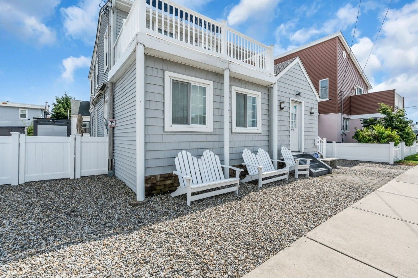 A two-story house with a balcony, a gravel driveway, and three Adirondack chairs.