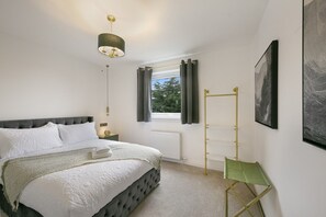 A bedroom with a bed, a window with curtains, a radiator, a green chair, and artwork on the wall.