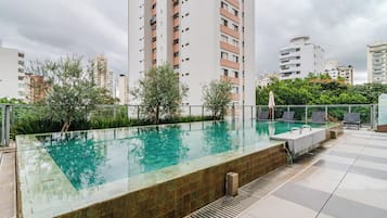Piscina no terraço com vista desobstruída para edifícios altos ao redor, cercada por vegetação e um deck revestido com azulejos.