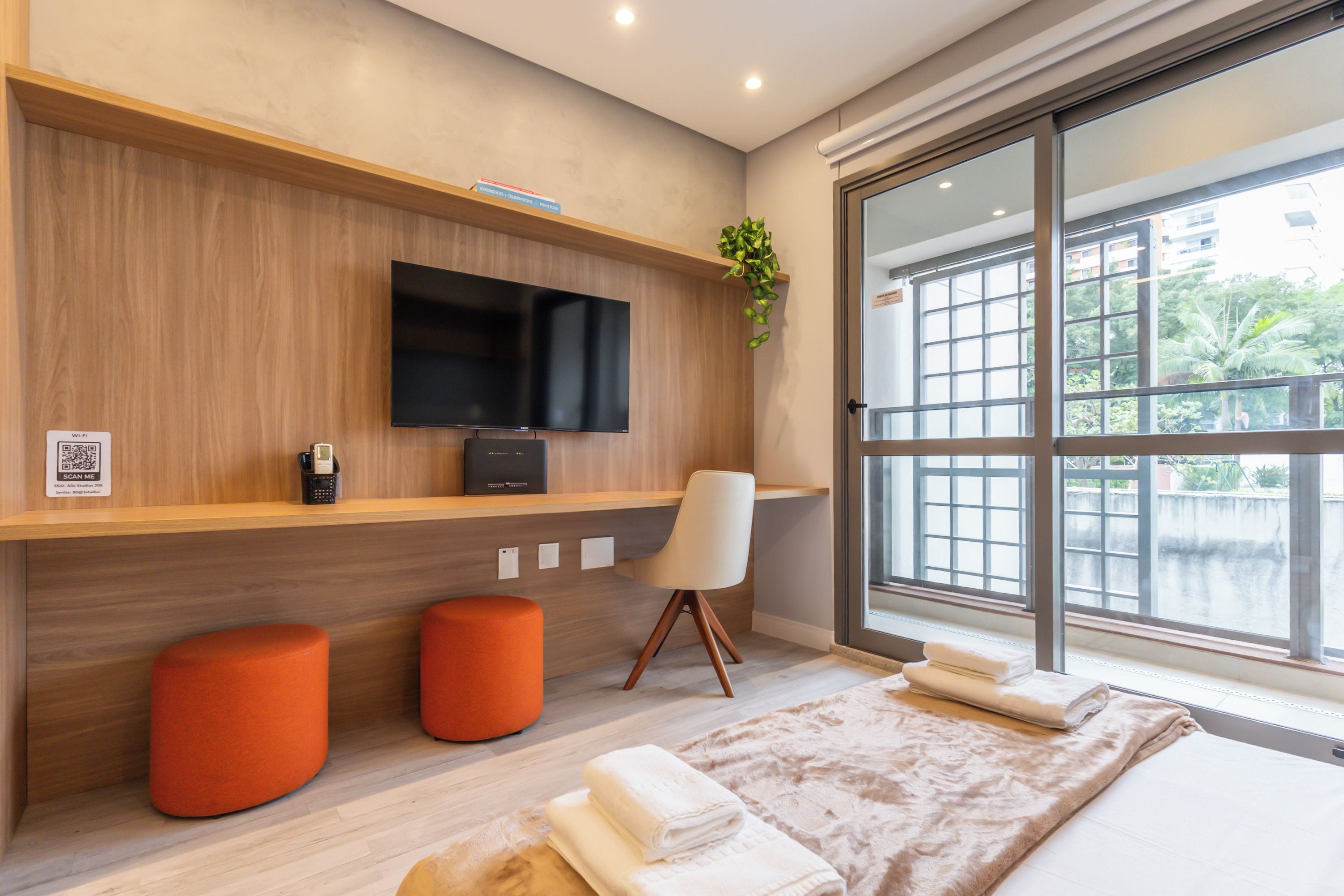 A modern hotel room with a large window, a flat-screen TV, a desk, two orange ottomans, and a bed with a fluffy blanket.