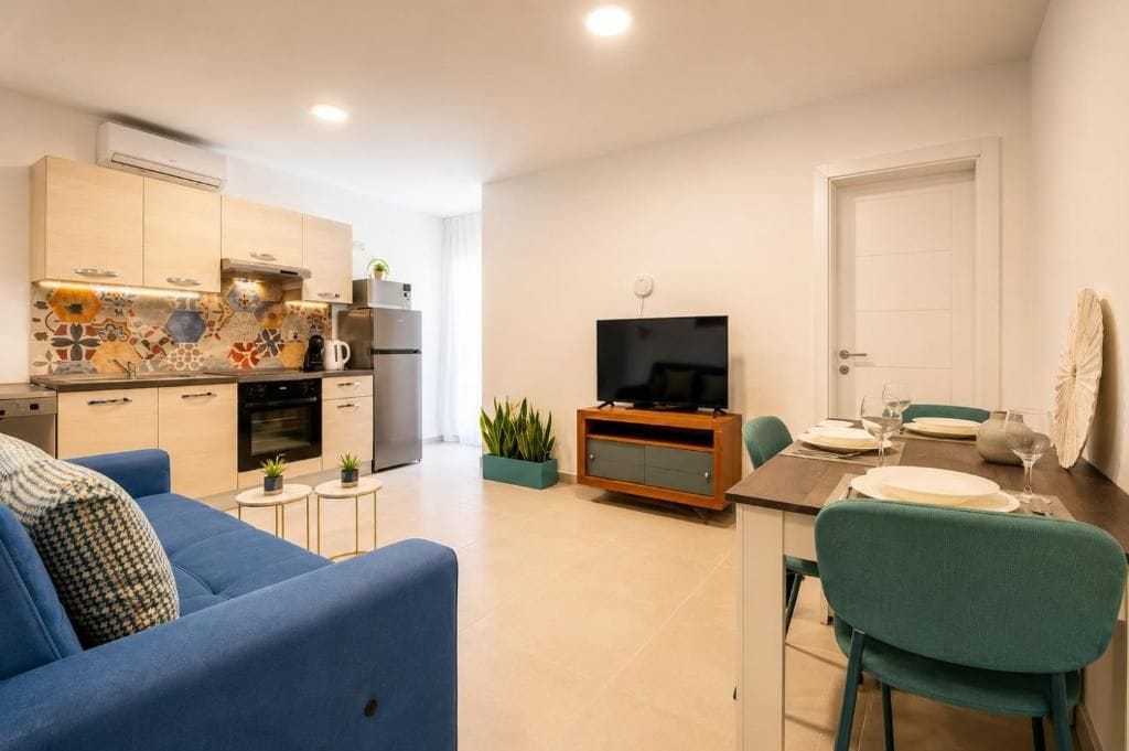 Una sala de estar moderna con un sofá azul, una mesa de comedor preparada para dos personas y una cocina con televisor y electrodomésticos.