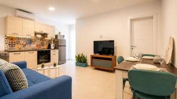 Una sala de estar moderna con un sofá azul, una mesa de comedor preparada para dos personas y una cocina con televisor y electrodomésticos.