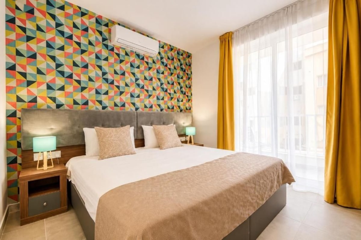 Quarto de hotel com parede estampada em padrão geométrico, uma cama grande com cabeceira, duas mesas de cabeceira com abajures e uma janela com cortinas.