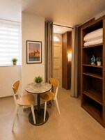 Una habitación con una mesa redonda, cuatro sillas, una planta en maceta, un cuadro enmarco, una estantería de madera y una puerta que da a otra habitación.
