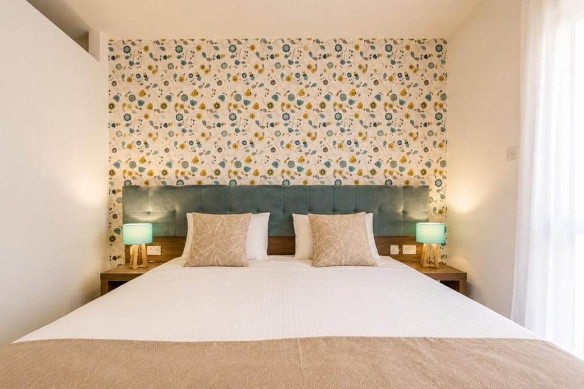 Quarto de hotel com uma cama grande, duas luminárias de cabeceira e cabeceira estampada com motivos florais.