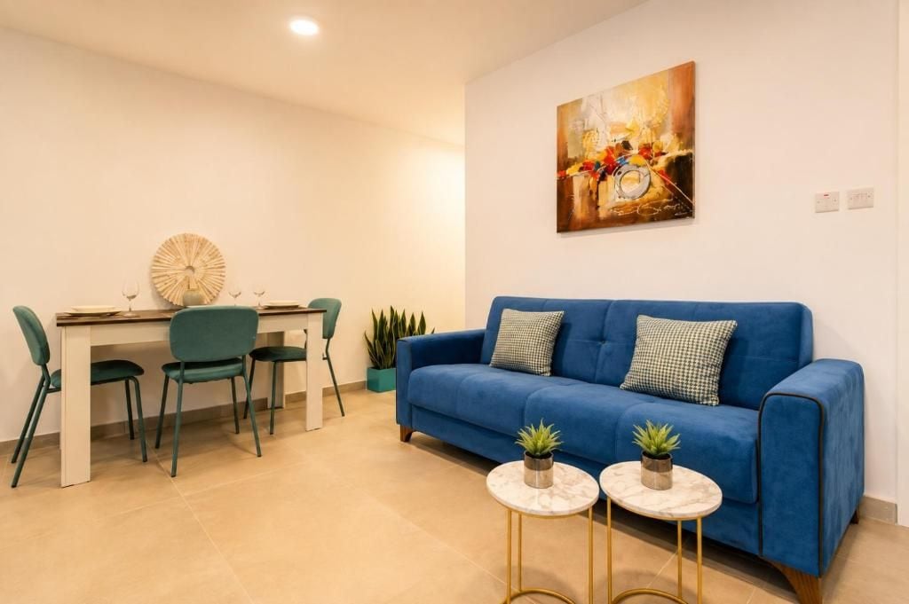 Una sala de estar con un sofá azul, dos mesitas redondas y un comedor con sillas y una mesa.