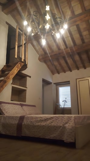 A room with a wooden staircase, a bed with a patterned bedspread, a chandelier, and a window with blinds. - Holiday Home 'Mare E Monti' with Mountain View, Private Pool, and Air Conditioning (Petriolo)