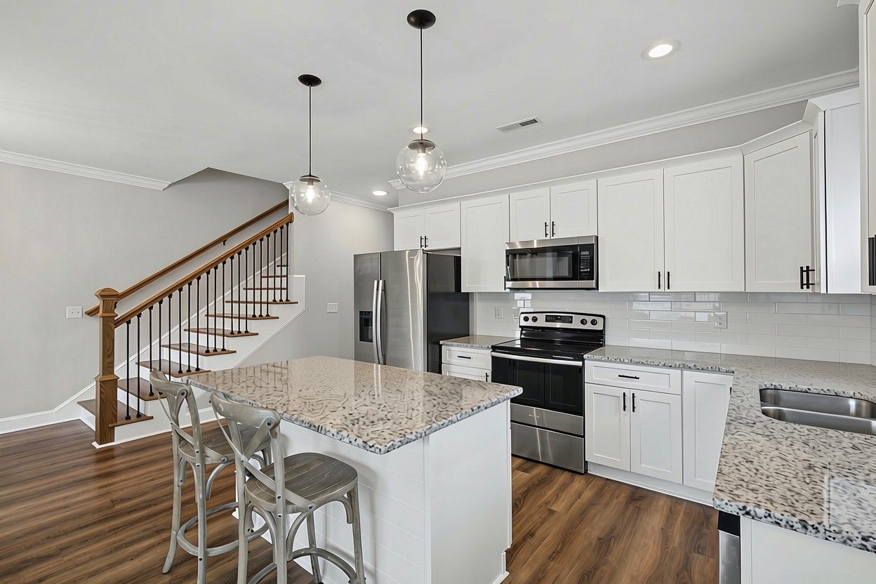 A modern kitchen with white cabinets, stainless steel appliances, a granite countertop island, and a wooden staircase.
