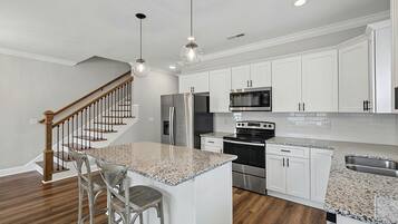 A modern kitchen with white cabinets, stainless steel appliances, a granite countertop island, and a wooden staircase.