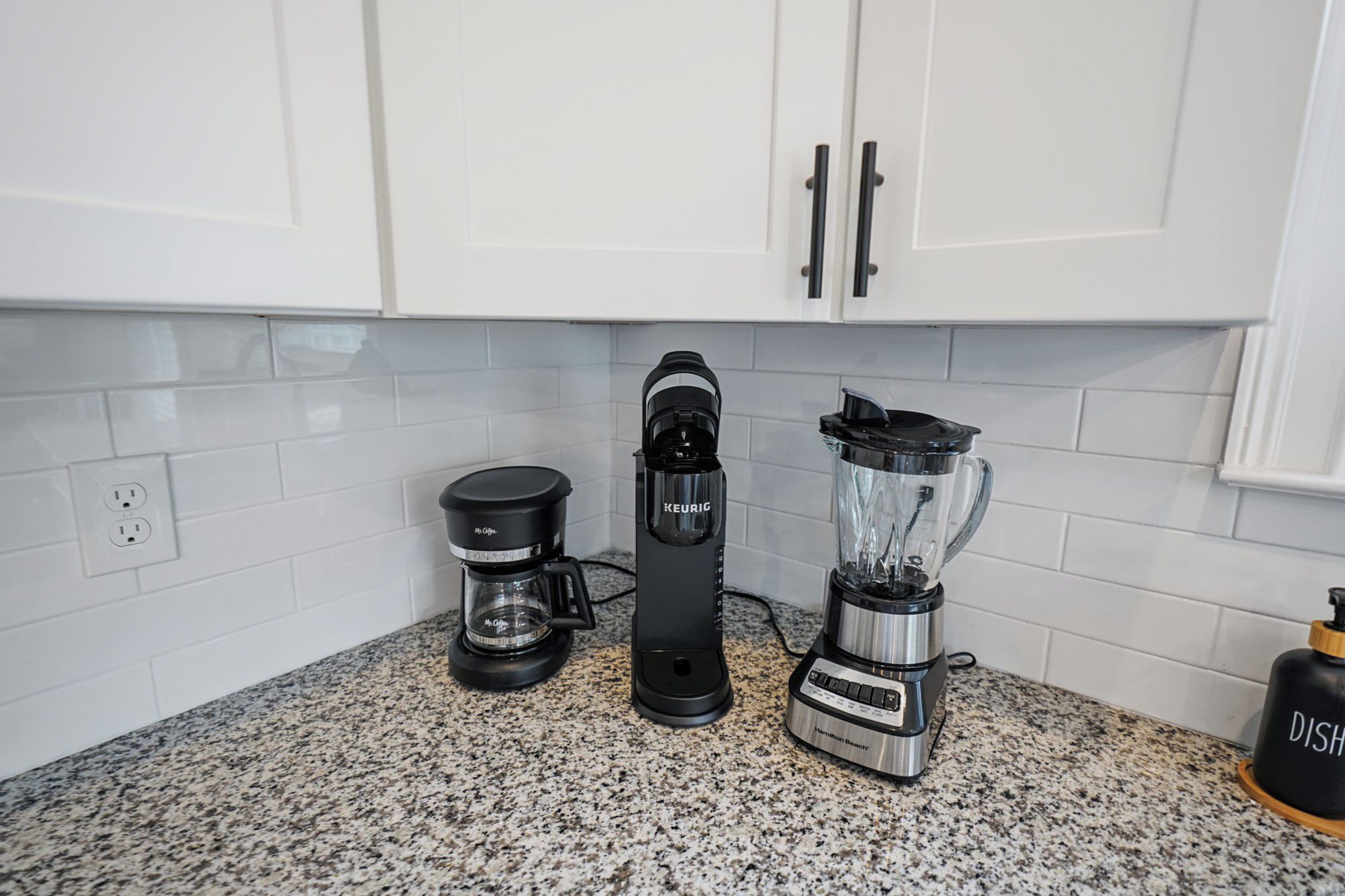 Ett kök med vita skåp, en kaffebryggare, en maskin för att brygga kaffe med kapslar (av märket Keurig) och en mixer från Hamilton Beach.
