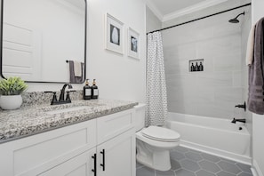 Une salle de bain moderne avec un comptoir en granit, des toilettes blanches, une douche dotée d’un rideau à motifs noirs et blancs, et une petite étagère contenant des articles de toilette.