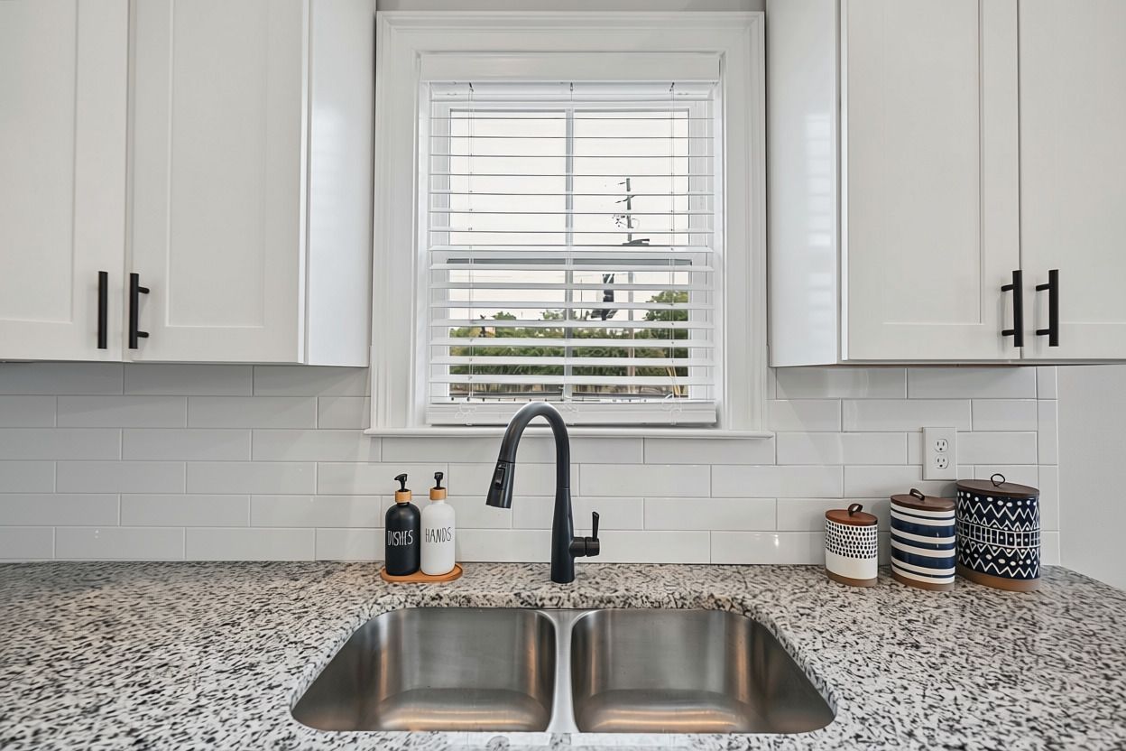 A kitchen with white cabinets, a double sink, a window with blinds, and decorative containers on the countertop.