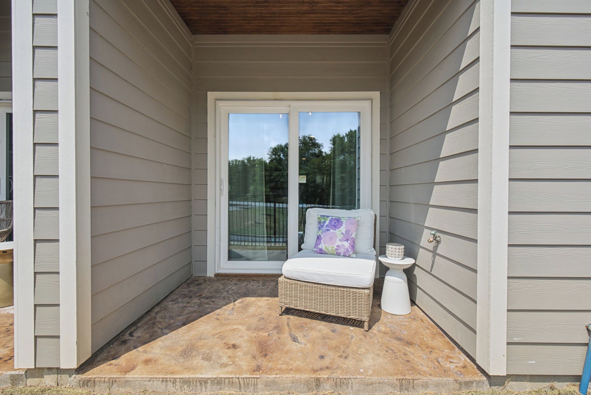 A covered porch with a wicker chair and a small table, a sliding glass door leading to a backyard, and a white exterior wall.