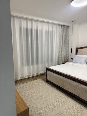 A bedroom with a bed, curtains, a rug, and a bedside table. - 2-bedroom with Extra Room in Nairobi,Westlands Near Sarit, with pool, gym, cafes (Nairobi)