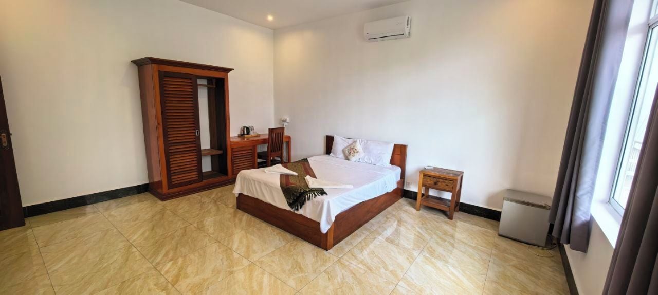 A hotel room with a bed, a wooden wardrobe, a desk, and a small bedside table.