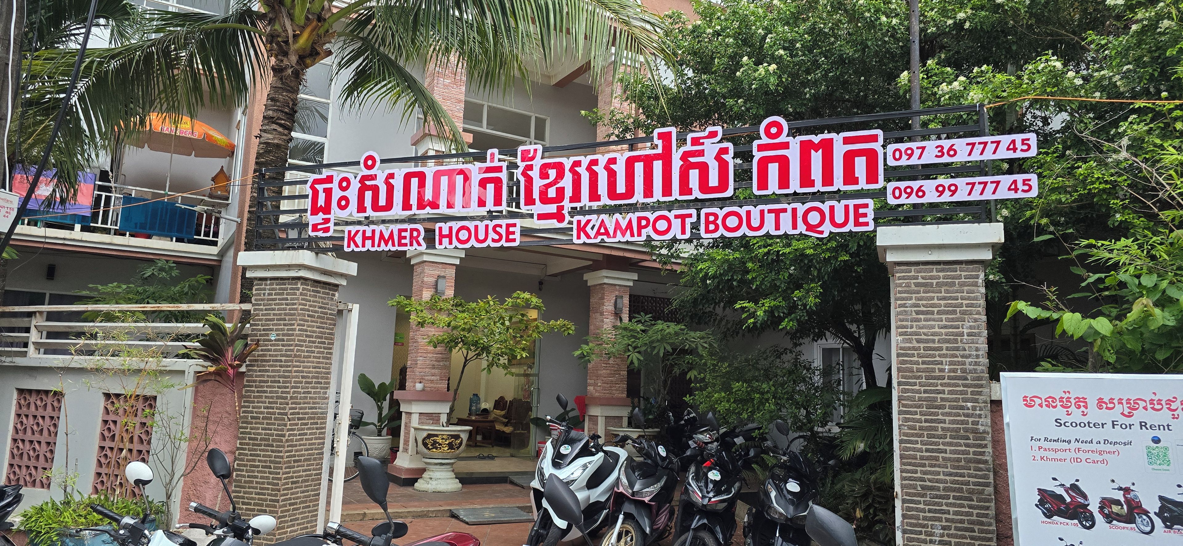 Un bâtiment portant une enseigne indiquant « KHMER HOUSE KAMPOT BOUTIQUE » et un panneau publicitaire pour la location de scooters, précisant qu’un passeport et une pièce d’identité sont requis.