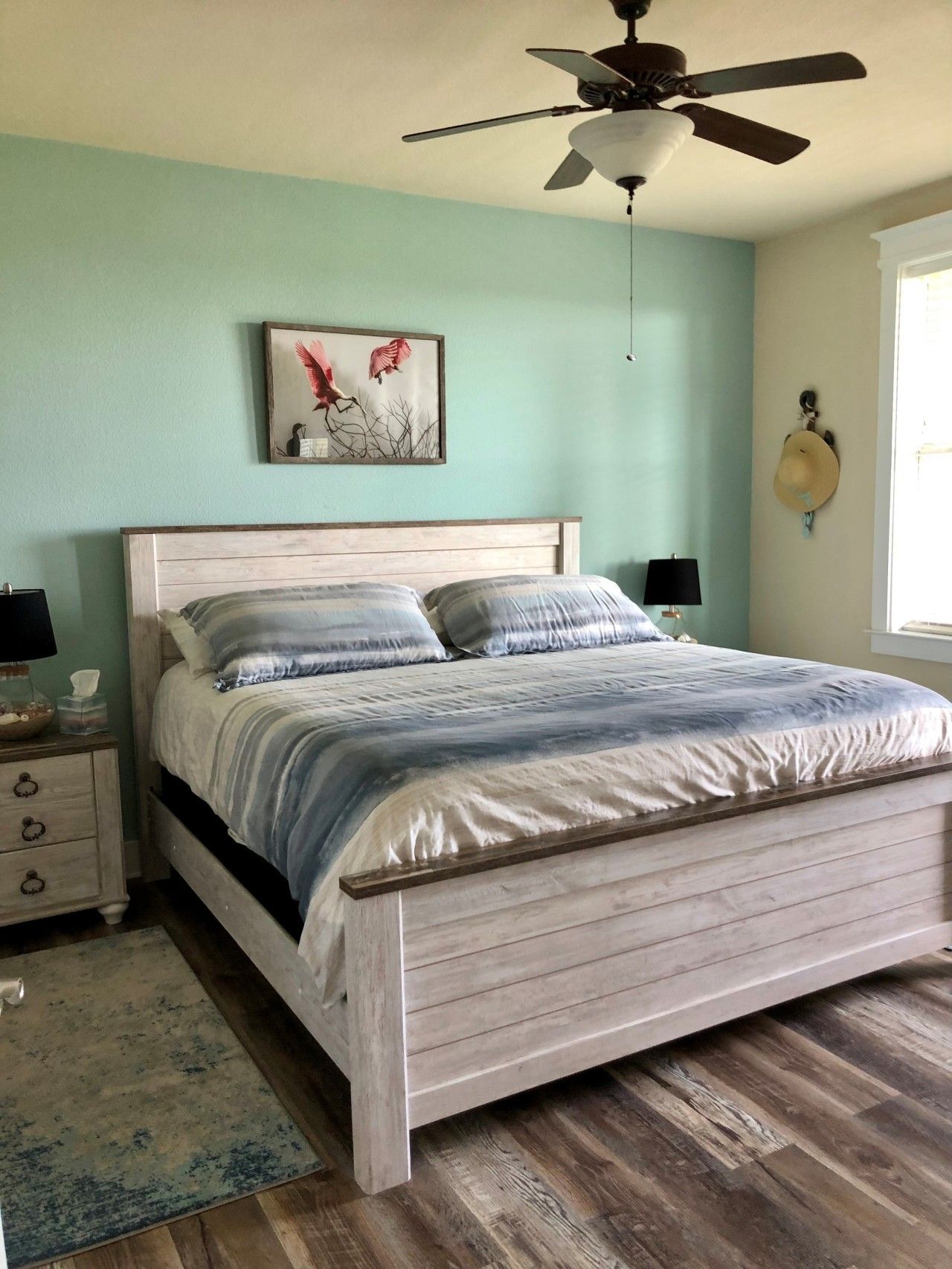 A bedroom with a bed, a ceiling fan, a nightstand, a lamp, and a wall-mounted hat.