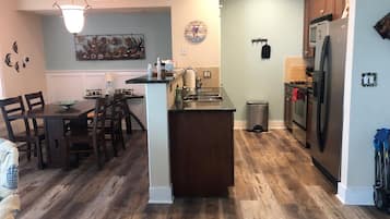 A kitchen with a dining area, wooden flooring, a refrigerator, and a clock on the wall.