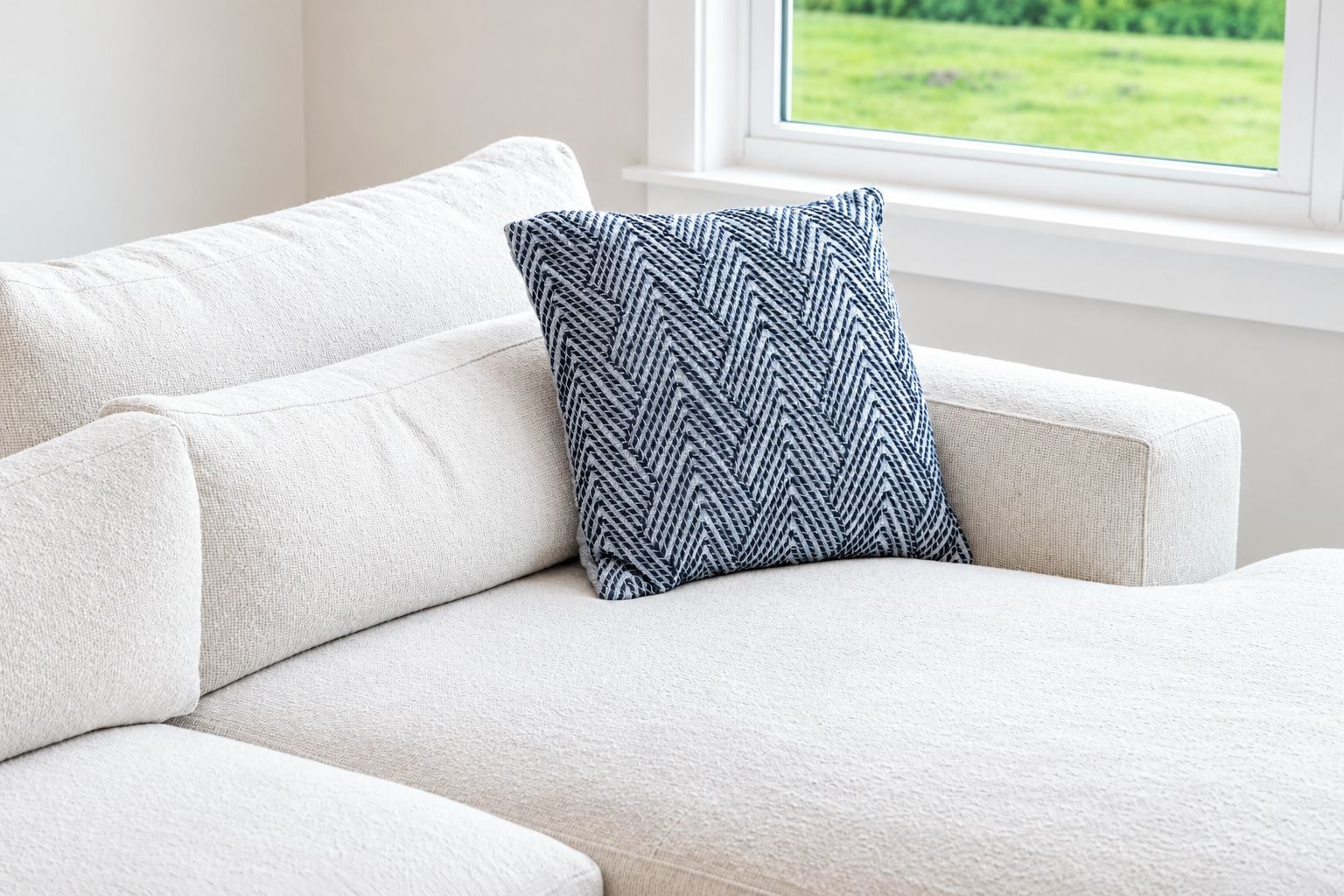 A white sofa with a patterned pillow in front of a window with a view of a green field.