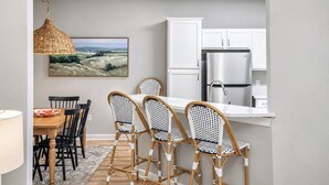 A modern kitchen with white cabinets, a stainless steel refrigerator, and a dining area with a wooden table and wicker chairs. - The Rebel Landing, Pool+3BR by Velvet Ditch Villas (Oxford)