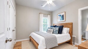 A bedroom with a bed, a ceiling fan, a window with curtains, a framed picture on the wall, and a door with a handle. - The Rebel Landing, Pool+3BR by Velvet Ditch Villas (Oxford)
