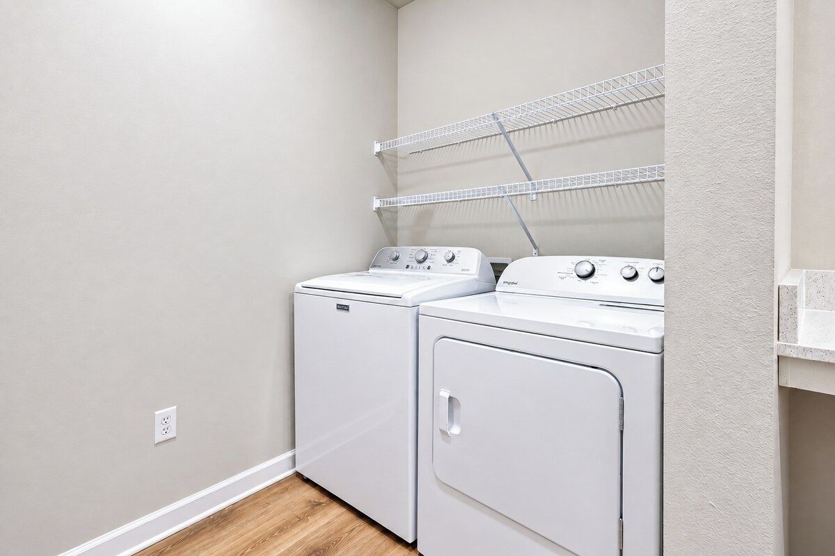 Une buanderie avec deux machines à laver et deux sèche-linge blancs, une étagère en métal au-dessus des sèche-linge et un mur blanc.
