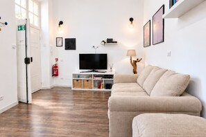 Un salon comprenant un canapé, un téléviseur à écran plat, une bibliothèque et une lampe fixée au mur.