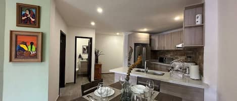 A dining area with a table set for four, a kitchen in the background, and artwork on the walls.