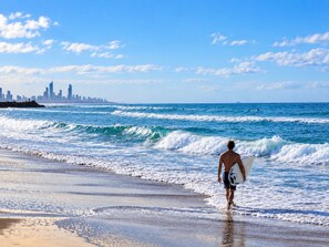 A beach scene with a surfer walking towards the ocean carrying a surfboard. - Palm and Sea Walkable Beachside Stay with Pool and Spa (Palm Beach)