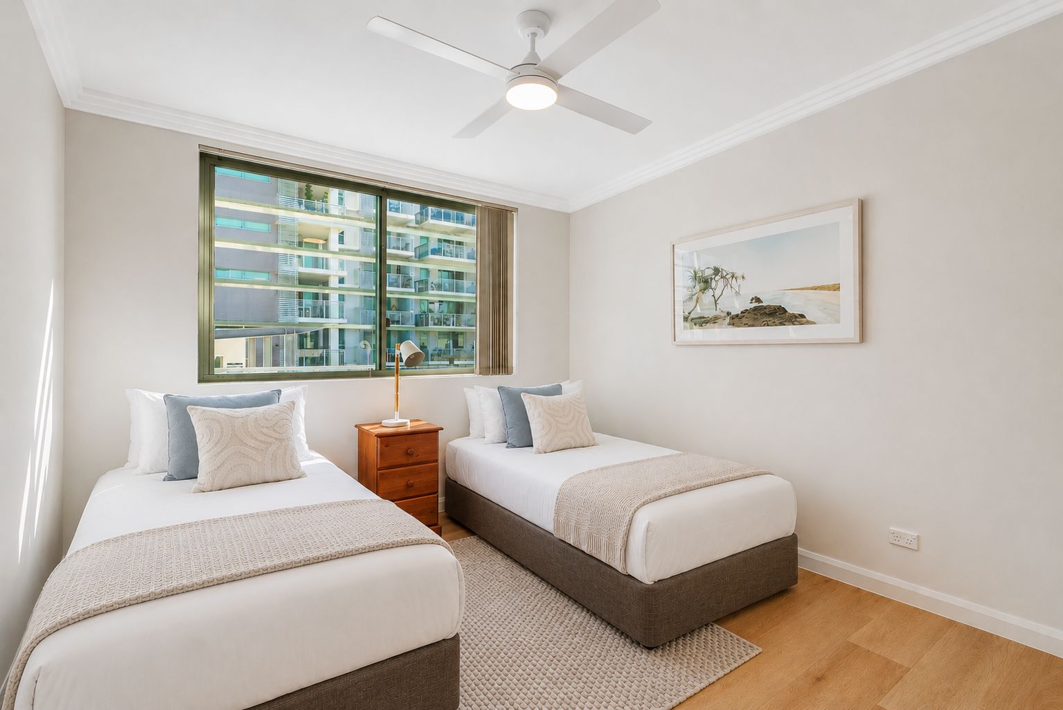 A bedroom with two beds, a ceiling fan, a window with a view of buildings, and a framed picture on the wall.