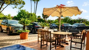 Área de varanda externa com piso de azulejos, móveis de madeira e um grande guarda-sol para proteção solar.