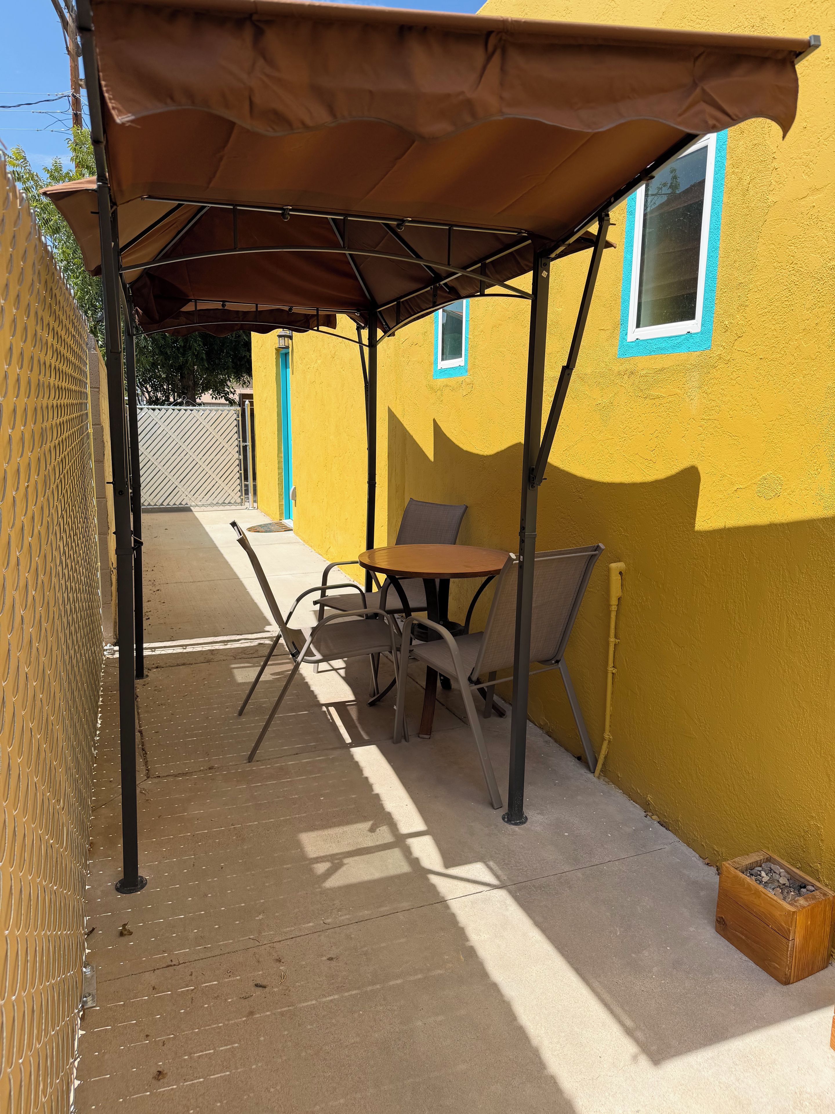 A patio area with a table and chairs under a canopy, adjacent to a yellow building with a window.