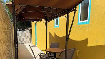 A patio area with a table and chairs under a canopy, adjacent to a yellow building with a window.