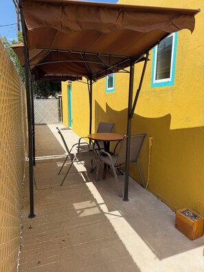 Eine Terrasse mit Tisch und Stühlen unter einem Baldachin, direkt neben einem gelben Gebäude mit einem Fenster.