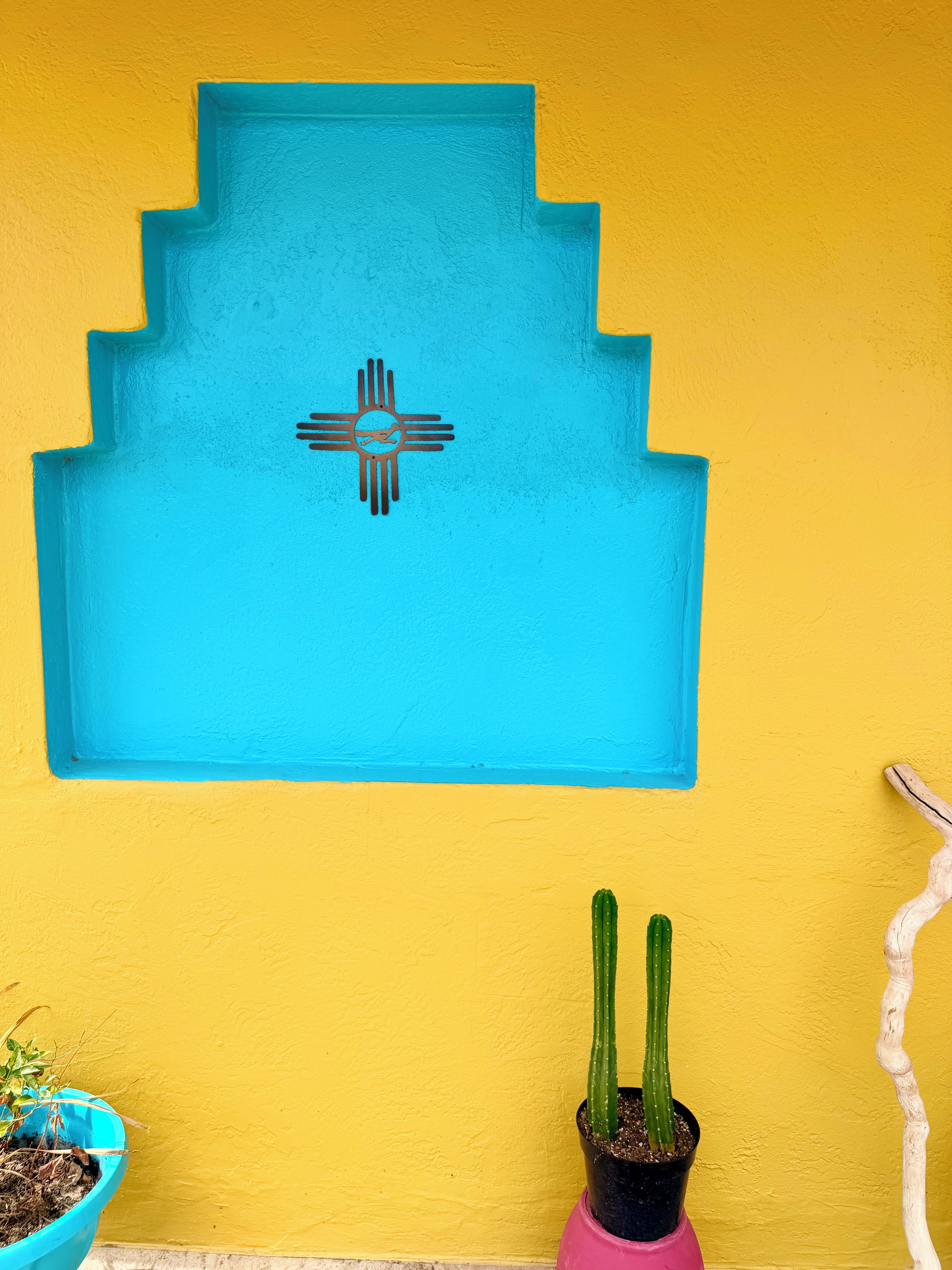 Eine Wand mit einem blauen geometrischen Muster und einem indianischen Symbol, eine Kakteepflanze in einem rosa Topf und ein hölzernes Deko-Objekt.