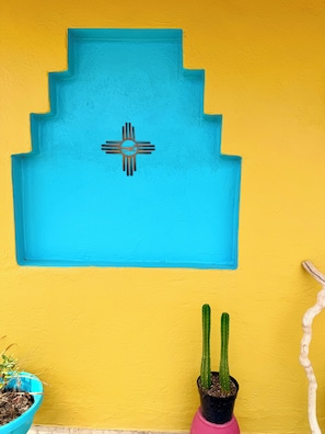 Eine Wand mit einem blauen geometrischen Muster und einem indianischen Symbol, eine Kakteepflanze in einem rosa Topf und ein hölzernes Deko-Objekt.