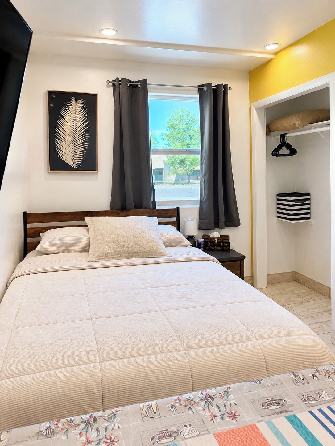 Un dormitorio con cama, mesita de noche, ventana con cortinas, un cuadro en la pared y un armario con perchero.