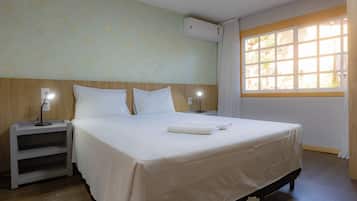 A hotel room with a bed, two bedside tables with lamps, a window with curtains, and an air conditioning unit on the wall.