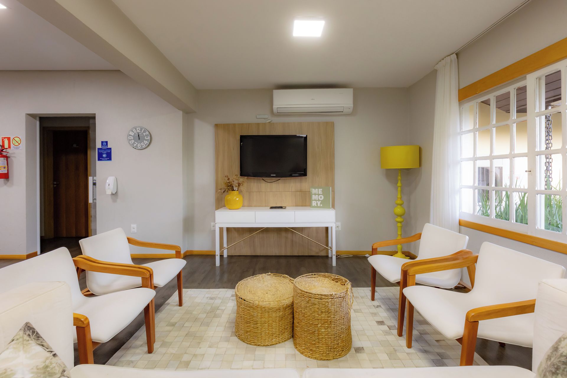 A hotel room with a white desk, two chairs, a TV, a lamp, and a window with curtains.