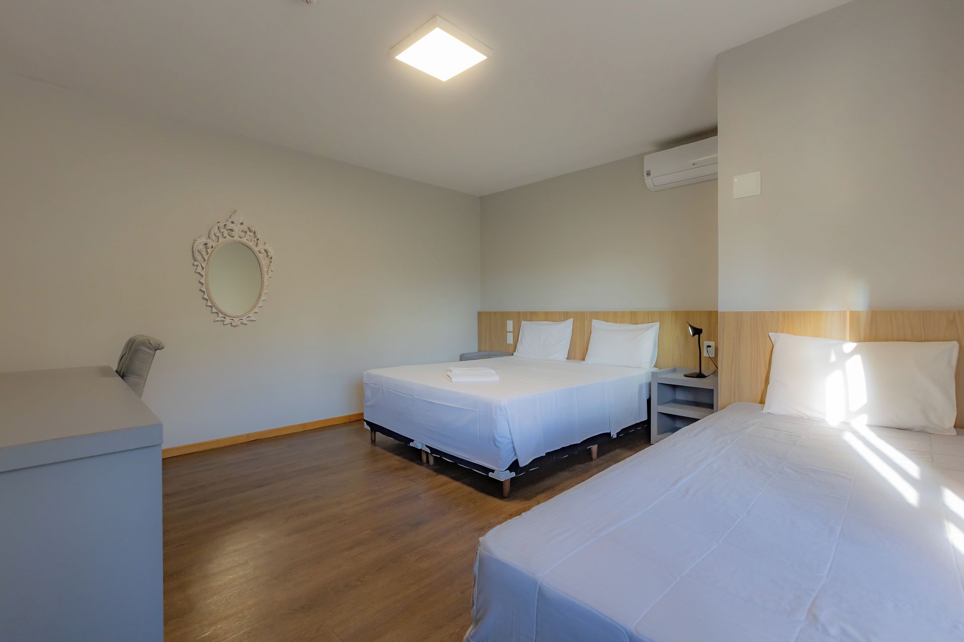 A hotel room with two beds, a bedside table with a lamp, a mirror on the wall, and an air conditioning unit.