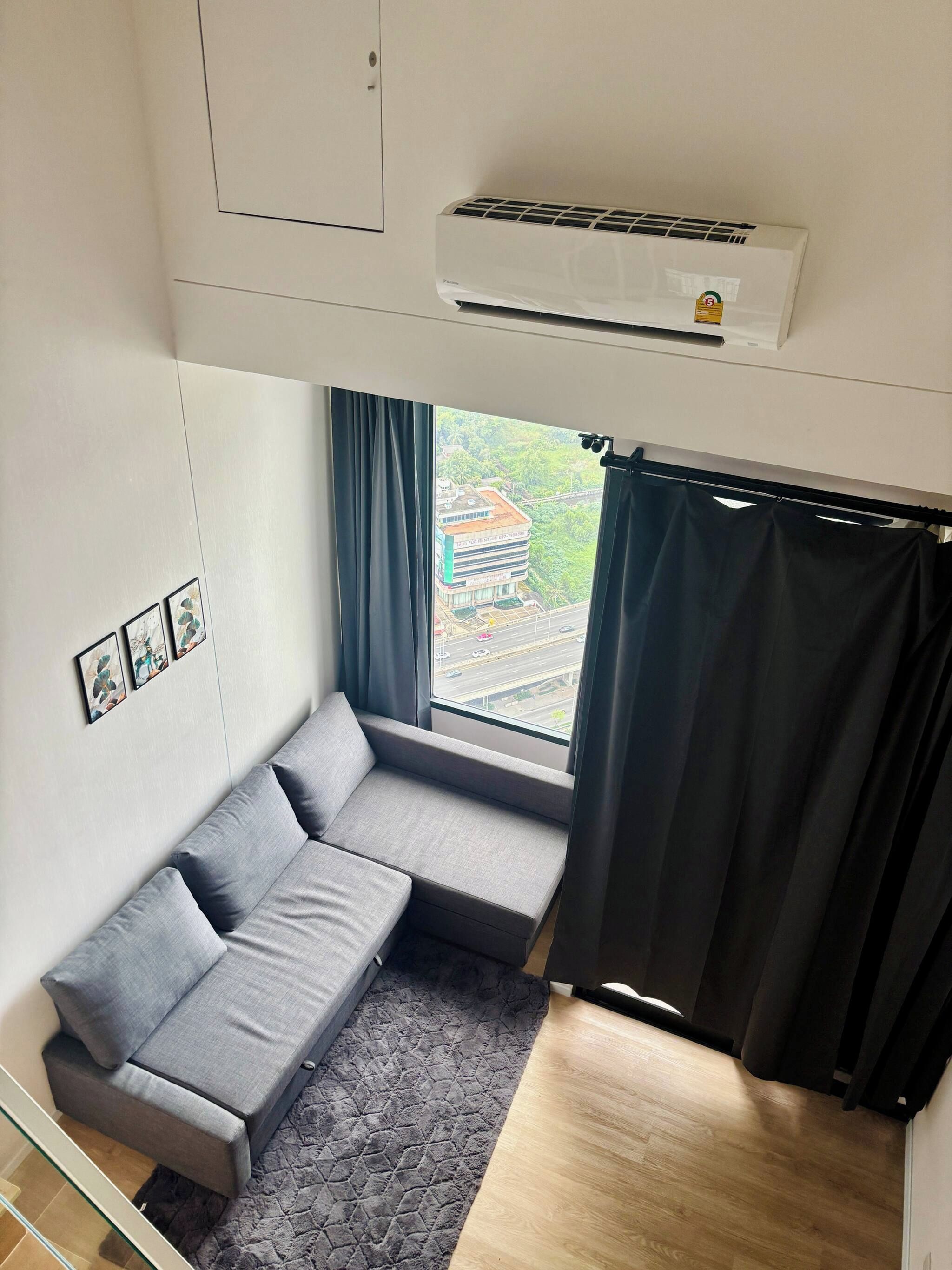 Una habitación compacta con un sofá gris, una ventana con cortinas, un aire acondicionado de pared y una pequeña alfombra.