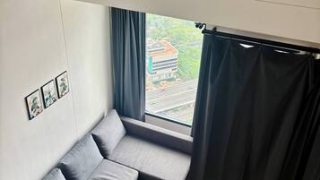 Quarto compacto com sofá cinza, janela com cortinas, ar condicionado fixado na parede e tapete pequeno.