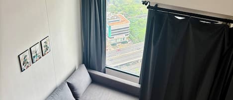 Una habitación compacta con un sofá gris, una ventana con cortinas, un aire acondicionado de pared y una pequeña alfombra.