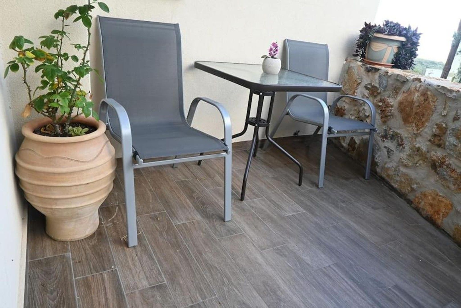 Un patio con dos sillas, una mesa, una planta en maceta y un muro de piedra.