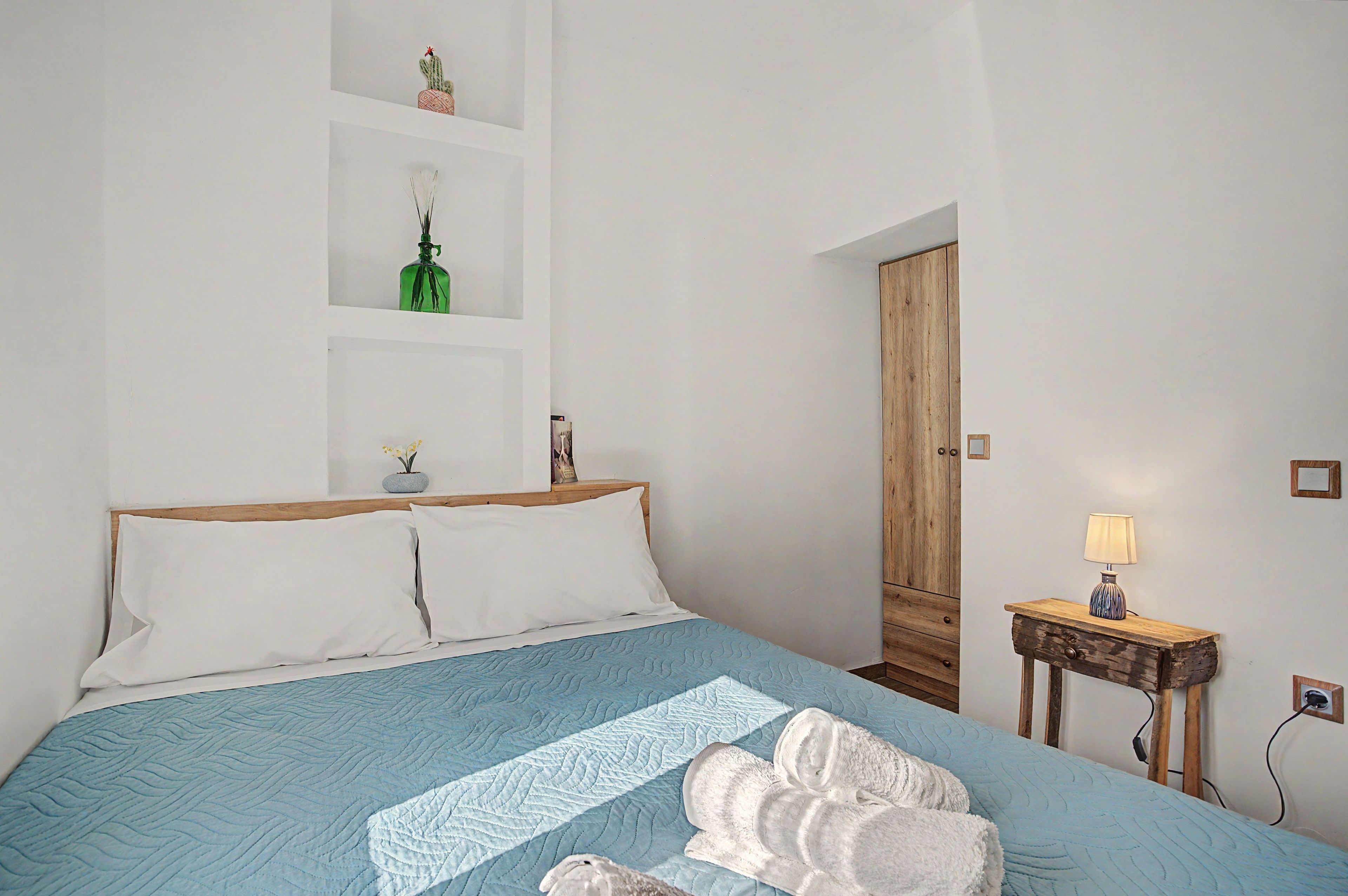 A bedroom with a bed, pillows, a bedside table with a lamp, a wooden wardrobe, and a shelf with decorative items.