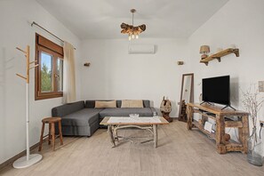 A modern living room with a sectional sofa, a coffee table, a TV on a wooden stand, and a large window with curtains. - Anezina – Cretan Hospitality in Agriana, near Analipsi Beach (Agriana)