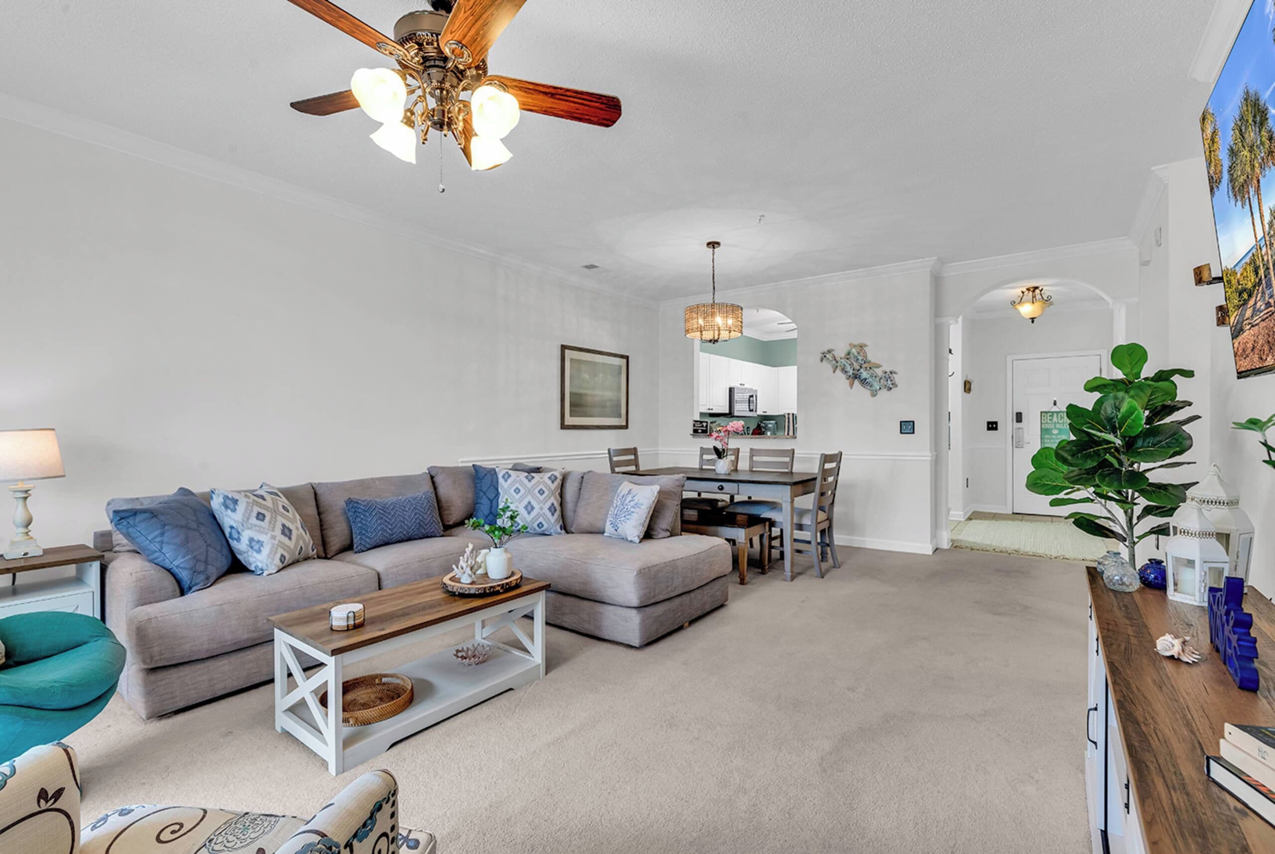 A living room with a ceiling fan, a dining area with a table and chairs, and a kitchen in the background.