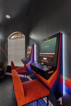 A room with two arcade gaming stations, a window with blinds, and a red chair. - New Luxe 5BR Resort | Pool, Pickleball | Theater (Mesa)