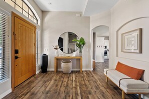 Un couloir avec une porte en bois, un miroir rond, une console ornée d’objets décoratifs, un panier en osier et un canapé avec un coussin orange.