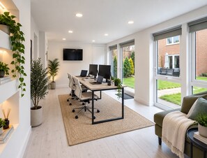 Ein moderner Arbeitsraum mit einem Schreibtisch, Stühlen, Computern und einem Fernseher. Pflanzen und ein Blick auf einen Garten sind vorhanden.